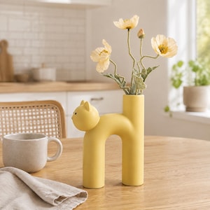 Può includere: Un vaso in ceramica giallo opaco a forma di gatto stilizzato con la schiena arcuata. Il vaso presenta un'apertura cilindrica per i fiori e una piccola testa di gatto con tratti del viso semplici. All'interno ci sono tre papaveri artificiali giallo pallido.