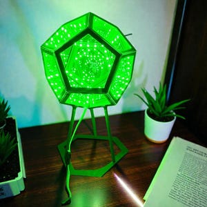 Può includere: Una lampada a specchio infinito a forma di dodecaedro che emette una luce verde brillante. La struttura geometrica presenta molteplici facce pentagonali con luci LED interne che creano un effetto tunnel ripetitivo, su un supporto in metallo verde.