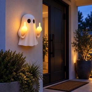 Op de afbeelding: Een witte, aan de muur gemonteerde spookvormige lamp met twee zwarte ovale ogen. De spookfiguur heeft twee armen omhoog, waarbij elke hand een middelvinger opsteekt. Het licht verspreidt een warme, zachte gloed vanuit het lichaam en de handen.