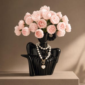 Peut inclure: Un vase en céramique noire brillante en forme de buste humain, rempli d'un grand bouquet de pivoines rose pâle. Un collier de perles blanches à deux rangs avec un fermoir doré et un petit pendentif en forme de cœur orne le cou du vase.
