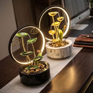 以下が含まれることがあります： 照明付きLEDリングを備えた2つの円形卓上噴水。1つは黒い大理石模様のベースに緑の蓮の葉の層があり、もう1つは白い大理石模様のベースに金色の蓮の葉の層が重なっています。