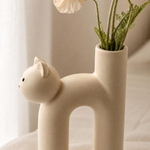May include: A matte beige ceramic vase shaped like a stylized cat with a smooth, arched body. The cat's head features small pointed ears and simple black dot eyes. A cylindrical opening for flowers extends upward from the cat's back section.