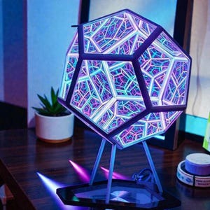 Peut inclure: Une lampe miroir à l'infini en forme de dodécaèdre avec un cadre sombre et des lumières LED internes créant un motif géométrique répétitif dans des tons de bleu, violet et rose. La lampe repose sur un fin support métallique à trois pieds.