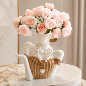 Peut inclure: Un vase buste en céramique blanche représentant le haut du torse et la tête d'une personne, portant un corsage texturé marron et un collier de perles à deux rangs avec un pendentif cœur doré. Le vase contient un bouquet de pivoines rose pâle.