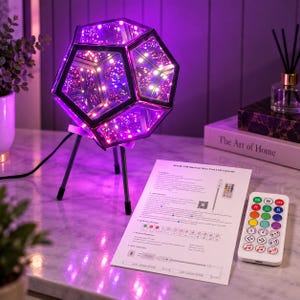 May include: A dodecahedron-shaped infinity mirror lamp with a black frame, emitting a vibrant purple LED light. The lamp stands on a three-legged metal tripod base and is accompanied by a white instruction sheet and a multi-button remote control.