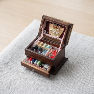 Op de afbeelding: Een kleine, open houten naaidoos met spoelen in verschillende kleuren, kleine scharen, naalden en andere naaibenodigdheden. De doos is gemaakt van donker hout met een gouden rand. De lade is ook open.