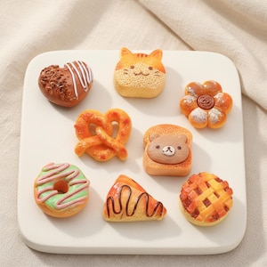 May include: A collection of miniature food items, including a heart-shaped pastry with chocolate sprinkles, a cat-shaped cake, a pretzel, a flower-shaped bread, a slice of pizza, a donut, a bear-shaped toast, and a pie. The items are arranged on a white square plate.