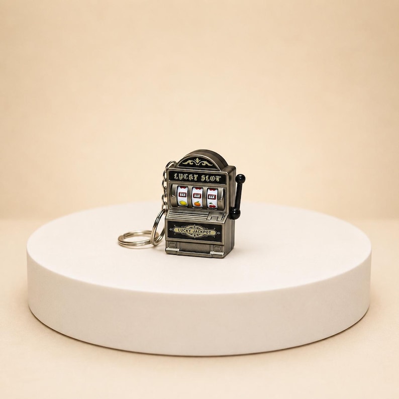 May include: A silver-toned metal keychain shaped like a miniature slot machine. The front features the text LUCKY SLOT above three spinning reels showing BAR and fruit symbols, with the text LUCKY JACKPOT below and a black side lever.