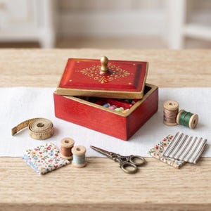 Op de afbeelding: Een rode houten naaidoos met een gouden decoratief ontwerp op het deksel, open om kleurrijke kralen te onthullen. Een meetlint, klosjes garen, stofresten en kleine scharen zijn rond de doos gerangschikt, wat wijst op een naai- of knutselthema.