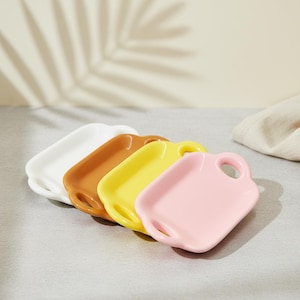 May include: Four small, rectangular ceramic trays in a row, each with a handle. The trays are white, brown, yellow, and pink. The trays are on a gray surface with a shadow of a leaf.