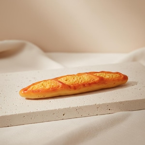 May include: A miniature baguette replica featuring a golden-brown, crispy-looking crust with distinct diagonal scoring marks along the top. The bread loaf has a realistic texture with a warm, toasted color palette and a slightly elongated, tapered shape.