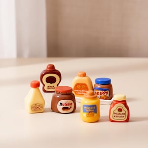 Pode incluir: Uma coleção de pequenas garrafas de condimentos, incluindo Nutella, mostarda e ketchup. As garrafas têm várias formas e cores, com rótulos como "Mustard Yellow" e "Tomato Ketchup". A disposição está em uma superfície clara.