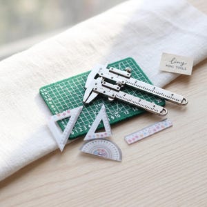 Puede incluir: Un juego en miniatura de herramientas de metal plateado, que incluye un calibrador, triángulos, un transportador y una regla, dispuestos sobre una esterilla de corte verde. El conjunto está etiquetado como "tiny MINI TOOLS". Las herramientas tienen marcas detalladas.