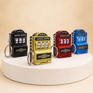 May include: Four miniature slot machine keychains in black, gold, red, and blue. Each keychain features a metal ring, three spinning reels with fruit and bar symbols, and the text Lucky Slot and Lucky Jackpot printed on the front surface.