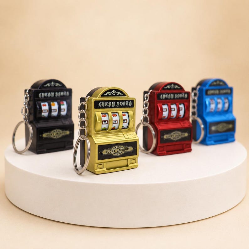 May include: Four miniature slot machine keychains in black, gold, red, and blue. Each keychain features a metal ring, three spinning reels with fruit and bar symbols, and the text Lucky Slot and Lucky Jackpot printed on the front surface.