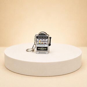 May include: A silver-toned metal slot machine keychain featuring a black lever on the right side. The front displays the text "LUCKY SLOTH" above three spinning reels showing fruit symbols, with the text "LUCKY JACKPOT" printed on the lower section.