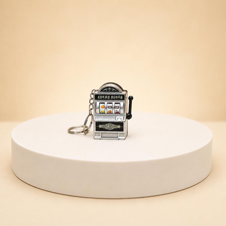 May include: A silver-toned metal slot machine keychain featuring a black lever on the right side. The front displays the text "LUCKY SLOTH" above three spinning reels showing fruit symbols, with the text "LUCKY JACKPOT" printed on the lower section.