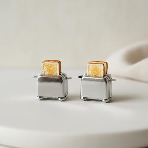 May include: Two miniature chrome toasters, each with two slices of toasted bread. The bread is golden brown, and the toasters have small levers and feet. The background is a soft, neutral color.