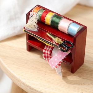 May include: A miniature wooden sewing station, stained a deep red, with spools of colourful thread, lace, scissors, and ribbons. The spools of thread are orange, red, yellow, green, pink, and blue. The station sits on a wooden surface.
