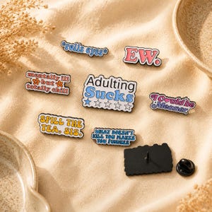 May include: A collection of seven enamel pins featuring various phrases including Adulting Sucks, EW, rolls eyes, mentally ill but totally chill, I could be meaner, Spill the tea sis, and What doesn't kill you makes you funnier, with one pin back shown.