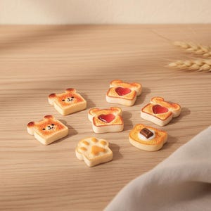 May include: A collection of miniature toast-shaped figurines with various designs. Some are shaped like bears, others have heart-shaped jelly, and one has a chocolate bar. The figurines are arranged on a light brown wooden surface.