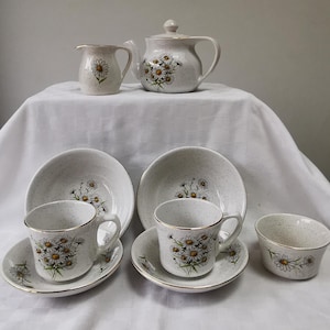 May include: A vintage ceramic tea set with a daisy floral pattern. The set includes a teapot, creamer, sugar bowl, two cups and saucers, and two bowls. The pieces are off-white with gold trim.