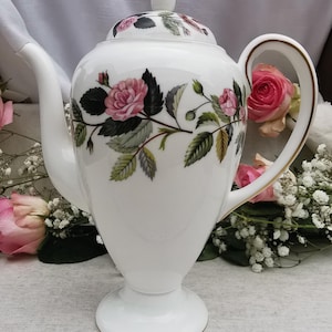Könnte beinhalten: Weiße Porzellan-Kaffeekanne mit Rosenmuster. Die Kanne hat einen gebogenen Ausguss, einen Griff mit Goldrand und einen passenden Deckel. Das florale Muster umfasst rosa Rosen, grüne Blätter und einen weißen Hintergrund. Die Kanne steht auf einem Sockel.