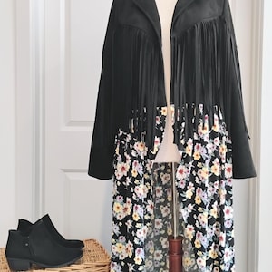 May include: A long-sleeved black jacket featuring a fringe detail across the chest and shoulders, paired with a black floral print skirt that has a high-low hemline. The floral pattern consists of yellow, white, and light purple flowers.