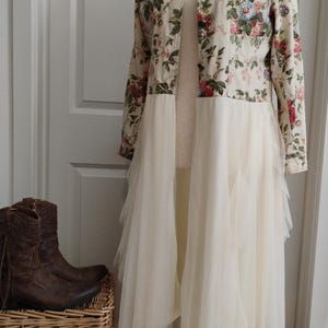 May include: A long-sleeved jacket featuring a cream base with a colorful floral print, button-down front, and a long, layered, sheer white tulle skirt attached at the waist. A pair of brown leather cowboy boots sits nearby.