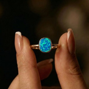 Puede incluir: Un anillo de oro con una gran ópalo de corte cuadrado en tonos azules y verdes vibrantes. El anillo se sostiene, mostrando el brillo de la piedra preciosa. La banda es de oro pulido.