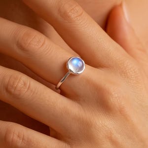 Pode incluir: Um anel de prata com uma pedra da lua redonda em corte cabochão, exibindo um brilho azul iridescente subtil. A gema está cravada num aro de prata lisa e polida sobre uma banda fina e simples, criando um visual limpo e elegante no dedo.