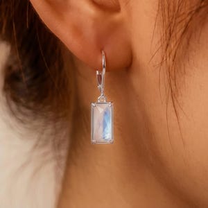 May include: A silver leverback earring featuring a rectangular, faceted moonstone with a translucent, iridescent blue sheen. The stone is set in a simple metal frame, hanging from a polished silver hook that secures to the earlobe.