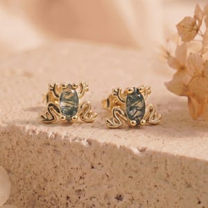 May include: A pair of gold-toned stud earrings shaped like frogs, each featuring an oval-cut moss agate gemstone set in the center. The metal frog design includes detailed legs and feet, with the green-patterned stone forming the body.