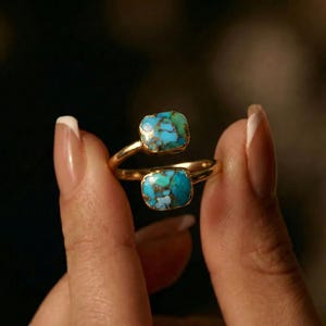 May include: A gold-toned ring featuring two square turquoise stones with a mix of blue, green, and brown hues. The ring's band curves around the finger, creating a unique and elegant design. The turquoise stones are the focal point of this jewelry piece.