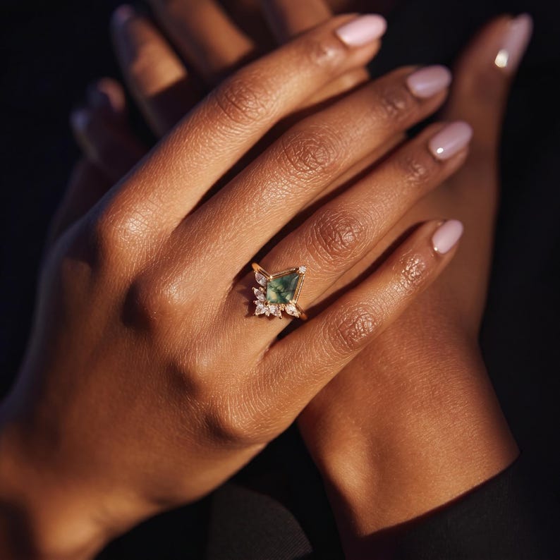 Puede incluir: Un anillo de oro con una gema verde en forma de cometa, rodeada de piedras blancas m&aacute;s peque&ntilde;as. El anillo est&aacute; en la mano de una persona, con los dedos y las u&ntilde;as visibles. El fondo es oscuro.