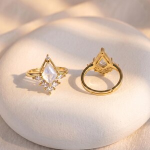 Puede incluir: Dos anillos dorados con un dise&ntilde;o geom&eacute;trico. Cada anillo presenta una piedra central en forma de diamante de color claro, rodeada de piedras m&aacute;s peque&ntilde;as y transparentes. Los anillos est&aacute;n sobre una superficie blanca texturizada.