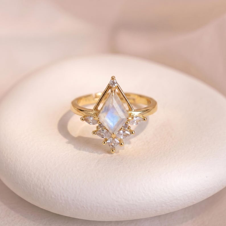 Puede incluir: Un anillo dorado con una piedra central iridiscente en forma de cometa. El anillo est&aacute; adornado con piedras preciosas m&aacute;s peque&ntilde;as y transparentes y un dise&ntilde;o geom&eacute;trico. El anillo est&aacute; sobre una superficie blanca texturizada.