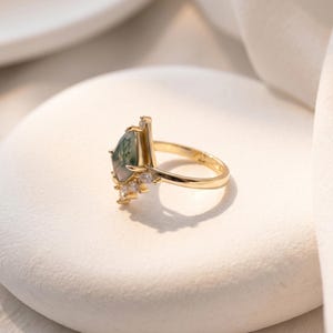 Puede incluir: Un anillo de oro con una piedra preciosa de &aacute;gata musgosa en forma de pera, con inclusiones verdes. El anillo tiene una banda delgada y est&aacute; adornado con peque&ntilde;as piedras preciosas transparentes. El anillo est&aacute; sobre una superficie blanca.
