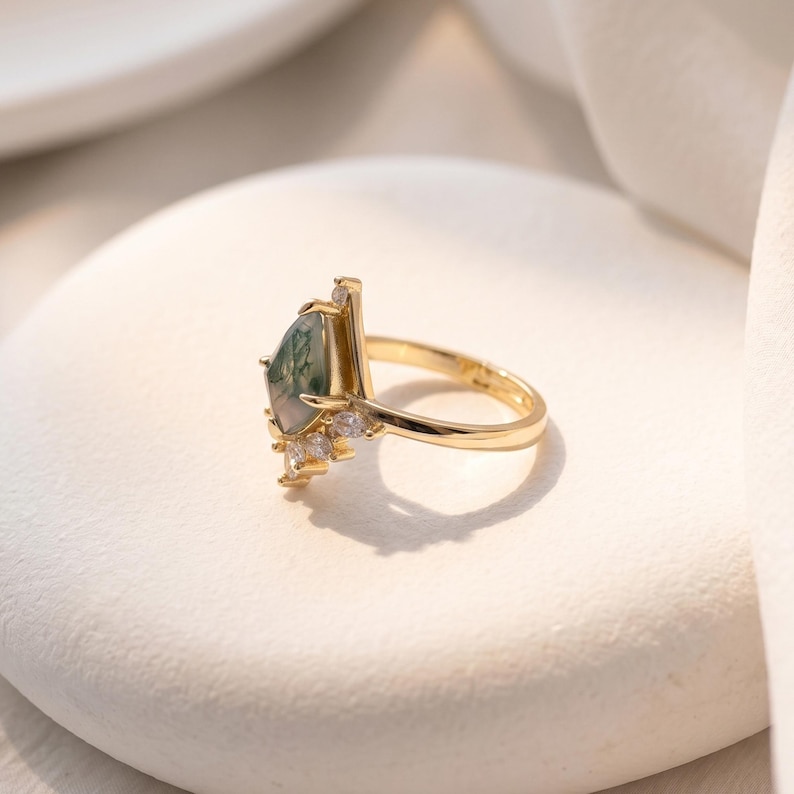 Puede incluir: Un anillo de oro con una piedra preciosa de &aacute;gata musgosa en forma de pera, con inclusiones verdes. El anillo tiene una banda delgada y est&aacute; adornado con peque&ntilde;as piedras preciosas transparentes. El anillo est&aacute; sobre una superficie blanca.