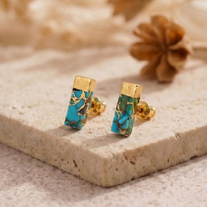 Pode incluir: Um par de brincos de tarracha com pedras retangulares de cor turquesa com veios castanhos e tampas de metal dourado. Cada brinco tem um fecho de pino, exibindo uma combinação de pedra azul brilhante e detalhes em metal dourado.