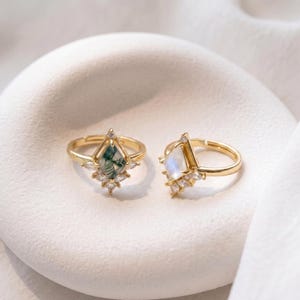 Puede incluir: Dos anillos dorados con piedras preciosas en forma de diamante. Uno presenta una piedra verde y blanca, el otro una piedra blanca. Ambos anillos est&aacute;n adornados con peque&ntilde;as piedras transparentes.
