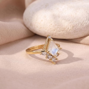 Puede incluir: Un anillo de oro con una piedra central de piedra lunar iridiscente en forma de cometa. El anillo presenta un grupo de piedras m&aacute;s peque&ntilde;as y transparentes y est&aacute; colocado sobre un fondo rosa suave. El dise&ntilde;o del anillo es elegante y moderno.