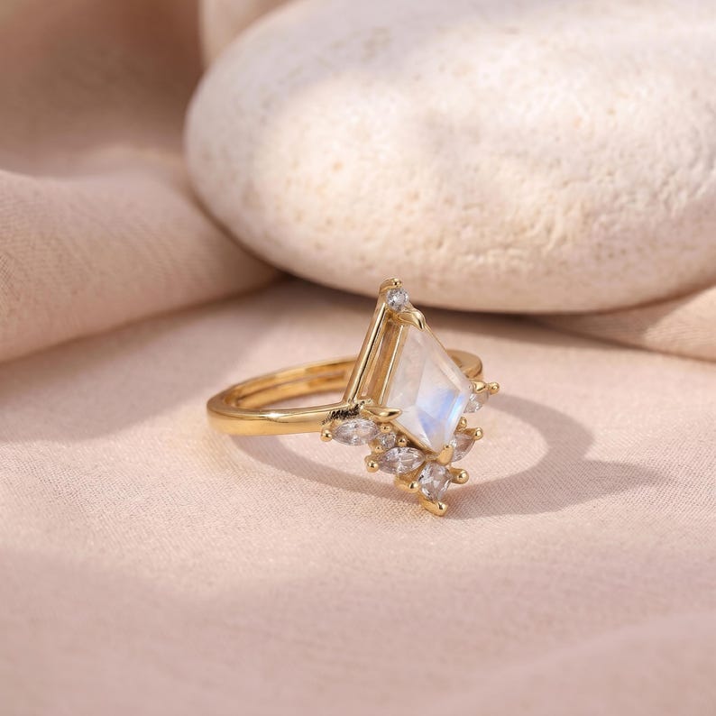 Puede incluir: Un anillo de oro con una piedra central de piedra lunar iridiscente en forma de cometa. El anillo presenta un grupo de piedras m&aacute;s peque&ntilde;as y transparentes y est&aacute; colocado sobre un fondo rosa suave. El dise&ntilde;o del anillo es elegante y moderno.