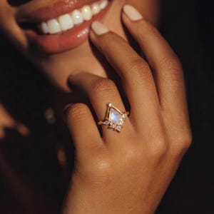 Puede incluir: Un anillo de oro con un dise&ntilde;o geom&eacute;trico que presenta una piedra preciosa azul claro central y piedras claras m&aacute;s peque&ntilde;as. El anillo est&aacute; en el dedo de una persona, con una sonrisa en el fondo.