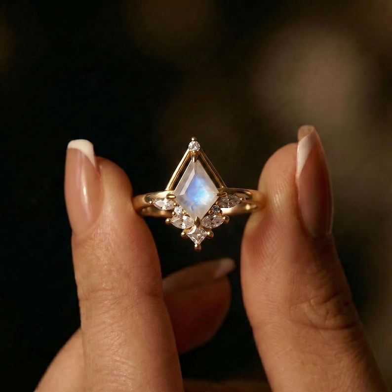 Puede incluir: Un anillo de oro con una piedra lunar iridiscente en forma de cometa. El anillo est&aacute; adornado con peque&ntilde;as piedras preciosas transparentes y tiene un dise&ntilde;o detallado. El anillo se sostiene contra un fondo borroso.