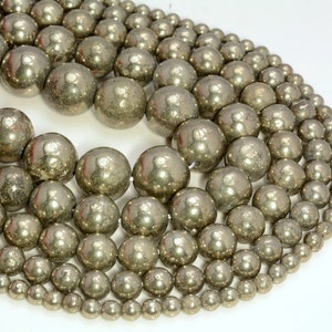 Puede incluir: Colección de cuentas de pirita metálicas de varios tamaños, dispuestas en forma curva. Las cuentas tienen un brillo reflectante plateado-dorado y están ensartadas, lo que sugiere suministros para joyería o artesanía.