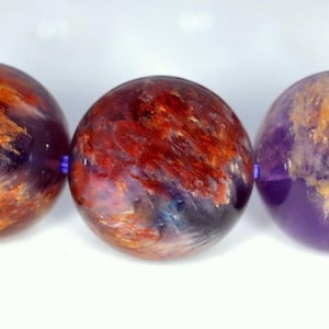 May include: Close-up of three round, multi-coloured gemstone beads. The beads are a mix of purple, orange, and red with a translucent, cloudy appearance.