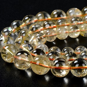 May include: Close-up of a strand of clear, round beads with a yellow-gold hue. Each bead has a reddish-orange band and internal golden striations. The beads are strung together, creating a jewelry item. The background is black.