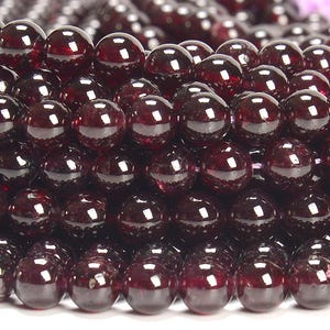 May include: Close-up of a pile of polished, round, dark red garnet beads. The beads are densely packed together, reflecting light and creating a glossy appearance. The beads are a deep, rich red color, with a smooth, spherical shape, perfect for jewelry making.