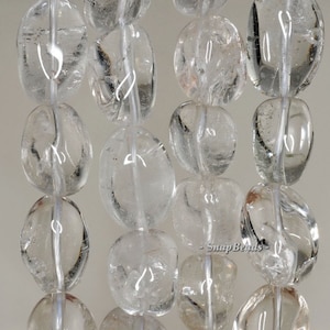 May include: A strand of clear quartz gemstone beads, each bead is oval shaped and has a smooth, polished finish. The beads are strung on a white thread.